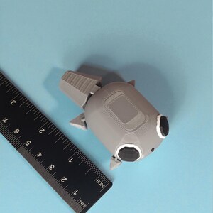 May include: A gray 3D-printed object with black accents, resembling a stylized robot or spacecraft, sits on a light blue surface. A black ruler with inch and millimeter markings is positioned nearby. The object's dimensions are approximately 3 inches in length.
