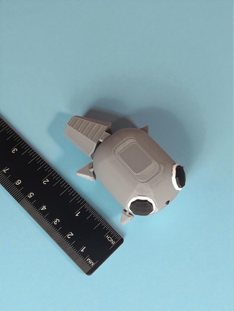 May include: A gray 3D-printed object with black accents, resembling a stylized robot or spacecraft, sits on a light blue surface. A black ruler with inch and millimeter markings is positioned nearby. The object's dimensions are approximately 3 inches in length.
