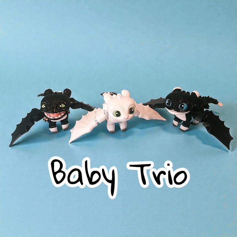 May include: Three toy dragons with outstretched wings are displayed against a blue background. Two are black with white accents, and one is white. The text "Baby Trio" is written in a white outlined black font.