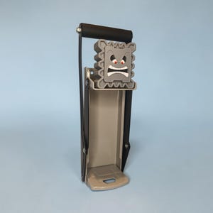May include: A gray can crusher with a black handle and a cartoonish, angry face design. The crusher is made of metal and plastic, with a lever for easy crushing. The design is reminiscent of a video game character.