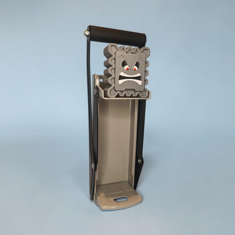 May include: A gray can crusher with a black handle and a cartoonish, angry face design. The crusher is made of metal and plastic, with a lever for easy crushing. The design is reminiscent of a video game character.