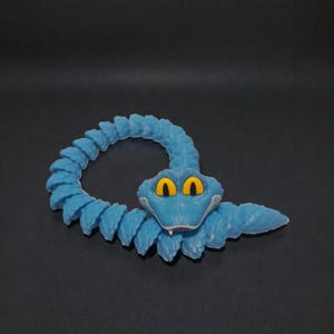 Puede incluir: Una serpiente de juguete azul con ojos amarillos y boca blanca. La serpiente est&aacute; enroscada en un bucle, con un cuerpo segmentado y una cola puntiaguda. El juguete es de pl&aacute;stico y est&aacute; sobre una superficie oscura.