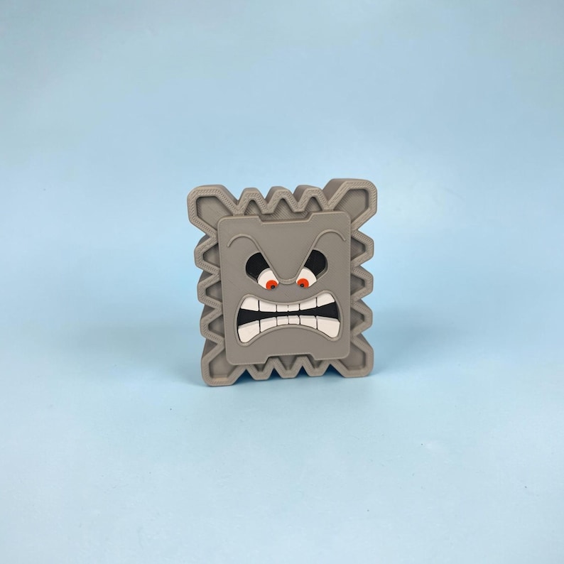 May include: A gray, square-shaped Thwomp figure with a detailed face, featuring angry red eyes, a wide open mouth with white teeth, and a jagged border. The figure is set against a light blue background.