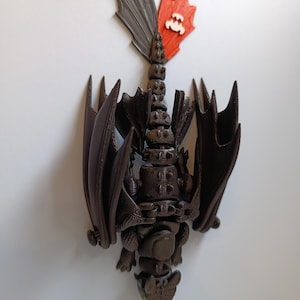 K&ouml;nnte beinhalten: Braune Drachenfigur mit ausgebreiteten Fl&uuml;geln und einem segmentierten K&ouml;rper. Der Drachenschwanz hat eine rote und graue Flosse mit einem wei&szlig;en Emblem. Die Figur ist eine detaillierte, dreidimensionale Darstellung eines Drachen.