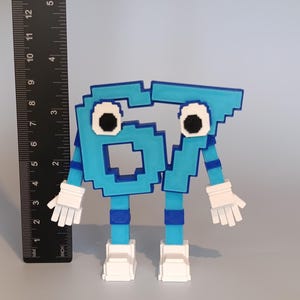 Pode incluir: Uma figura azul pixelizada com o n&uacute;mero 67, com luvas e sapatos brancos. A figura tem olhos pretos e brancos e mede aproximadamente 13 cm de altura. &Agrave; esquerda, h&aacute; uma r&eacute;gua preta com marca&ccedil;&otilde;es em polegadas e mil&iacute;metros.