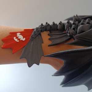 K&ouml;nnte beinhalten: Ein schwarzes und rotes Drachenfl&uuml;gel-Armband. Das Accessoire hat eine rote Flagge mit einem wei&szlig;en Emblem und schwarze Fl&uuml;geldarstellungen. Der Drachenfl&uuml;gel ist f&uuml;r das Tragen am Handgelenk konzipiert.