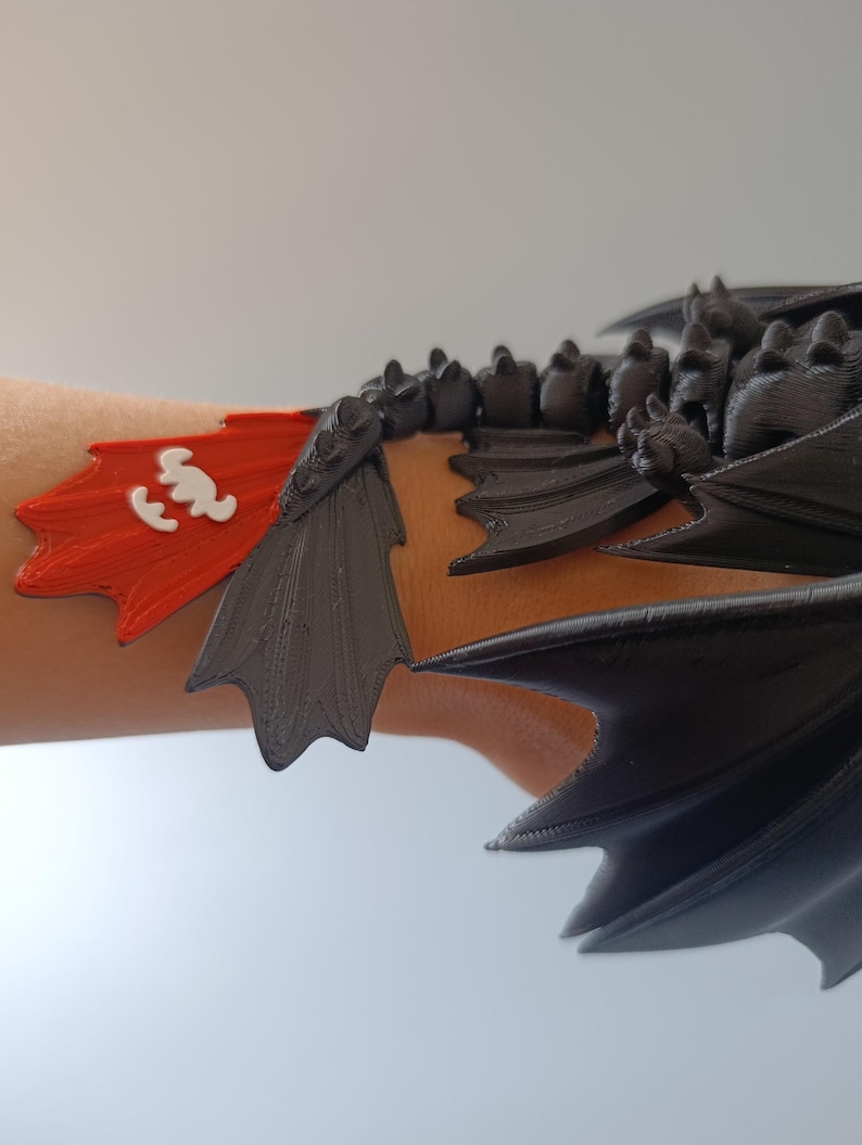 K&ouml;nnte beinhalten: Ein schwarzes und rotes Drachenfl&uuml;gel-Armband. Das Accessoire hat eine rote Flagge mit einem wei&szlig;en Emblem und schwarze Fl&uuml;geldarstellungen. Der Drachenfl&uuml;gel ist f&uuml;r das Tragen am Handgelenk konzipiert.
