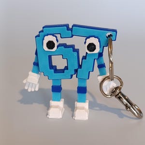 Pode incluir: Um chaveiro de figura pixelizada azul e branca com o n&uacute;mero 67. A figura tem bra&ccedil;os, pernas e p&eacute;s, e est&aacute; presa a um porta-chaves e fecho prateados. Os olhos da figura s&atilde;o pretos e brancos.