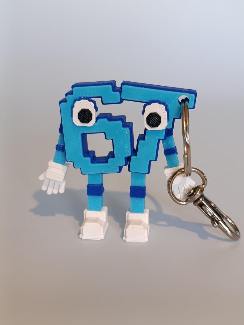 Pode incluir: Um chaveiro de figura pixelizada azul e branca com o n&uacute;mero 67. A figura tem bra&ccedil;os, pernas e p&eacute;s, e est&aacute; presa a um porta-chaves e fecho prateados. Os olhos da figura s&atilde;o pretos e brancos.