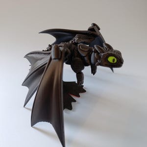K&ouml;nnte beinhalten: Eine schwarze Drachenfigur mit weit ausgebreiteten Fl&uuml;geln und detailliertem K&ouml;rper. Der Drache hat leuchtend gr&uuml;ne Augen und scheint zu fliegen. Die Figur steht vor einem schlichten wei&szlig;en Hintergrund.