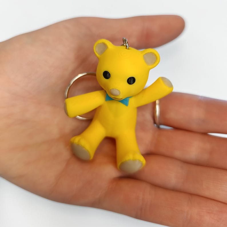 Puede incluir: Un llavero de oso amarillo con una peque&ntilde;a pajarita azul, ojos negros y una nariz de color claro. El oso tiene detalles claros en las orejas y las patas, y est&aacute; sujeto a una anilla met&aacute;lica circular para llaves, con un acabado amarillo brillante.