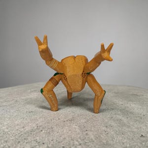 May include: A wooden figurine of a crab-like creature with its arms raised in a peace sign. The figurine is primarily tan with green and brown accents. The creature has three legs and a textured body.