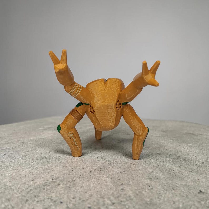 May include: A wooden figurine of a crab-like creature with its arms raised in a peace sign. The figurine is primarily tan with green and brown accents. The creature has three legs and a textured body.