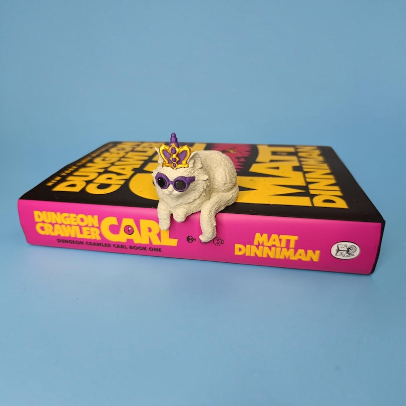 Op de afbeelding: Een wit kattenbeeldje met een paarse en gouden kroon en een paarse zonnebril, zittend op een boek getiteld Dungeon Crawler Carl van Matt Dinniman. Het boek heeft een zwarte kaft met opvallende gele en roze tekst en een roze rug.