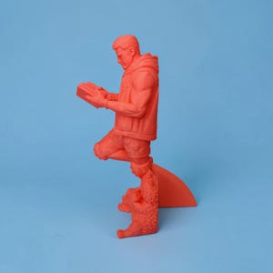 May include: A monochromatic coral-orange figurine depicting a person standing on one leg while reading a book. The figure wears a hooded jacket and shorts, with one foot resting on a textured, dragon-like creature that provides a base for the sculpture.