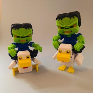 May include: Two toy figures of a green-skinned character with black hair and a blue shirt riding on white ducks with yellow beaks and feet. The figures have an angry expression.