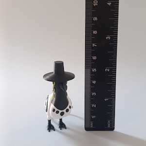 May include: A 3D-printed figurine of a bird wearing a black hat. The bird has a black beak and head, with a white body and black spots. The figurine is approximately 3 inches tall, as measured by the ruler in the image.