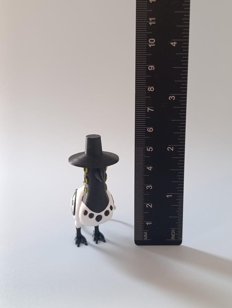 May include: A 3D-printed figurine of a bird wearing a black hat. The bird has a black beak and head, with a white body and black spots. The figurine is approximately 3 inches tall, as measured by the ruler in the image.