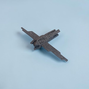 K&ouml;nnte beinhalten: Ein graues, lasergeschnittenes Modellflugzeug mit komplizierten geometrischen Mustern auf Fl&uuml;geln und Rumpf. Das Modell ist diagonal vor einem hellblauen Hintergrund positioniert und zeigt sein detailliertes Design und seine Handwerkskunst. Die Fl&uuml;gel des Flugzeugs sind ausgebreitet.