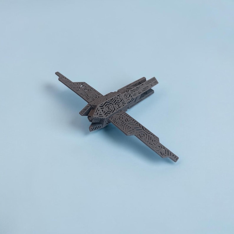 K&ouml;nnte beinhalten: Ein graues, lasergeschnittenes Modellflugzeug mit komplizierten geometrischen Mustern auf Fl&uuml;geln und Rumpf. Das Modell ist diagonal vor einem hellblauen Hintergrund positioniert und zeigt sein detailliertes Design und seine Handwerkskunst. Die Fl&uuml;gel des Flugzeugs sind ausgebreitet.