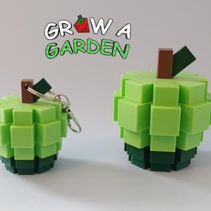 Puede incluir: Dos objetos verdes con forma de manzana pixelada, uno un llavero, el otro una pieza decorativa m&aacute;s grande. Ambos tienen tallos marrones y hojas verdes oscuras. El texto "GROW A GARDEN" est&aacute; encima en blanco y verde.