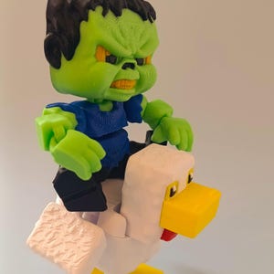 May include: A 3D-printed toy featuring a green monster figure with black hair and an angry expression, riding a white chicken with yellow beak and feet. The monster wears a blue shirt and black pants.