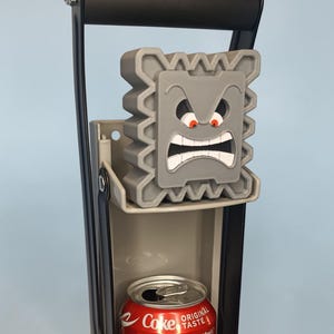 May include: A gray Thwomp-shaped can crusher with an angry face design. The crusher is mounted on a black and gray frame, with a crushed Coke can visible at the bottom. The design features red eyes and white teeth.