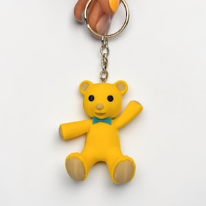Puede incluir: Un llavero de oso amarillo con una peque&ntilde;a pajarita de color verde azulado, ojos negros y nariz marr&oacute;n claro. El oso est&aacute; unido a una anilla de metal plateado mediante una cadena corta. Tiene un acabado liso con detalles en marr&oacute;n claro.