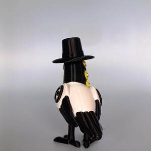 Puede incluir: Figura impresa en 3D de un p&aacute;jaro con sombrero de copa negro. El p&aacute;jaro tiene un cuerpo negro con detalles blancos en el pecho y las alas. Tiene ojos amarillos y se ve desde atr&aacute;s.