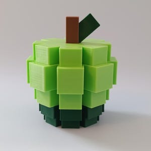Puede incluir: Una escultura de manzana impresa en 3D, compuesta por cubos verdes apilados, con un tallo marr&oacute;n y una hoja verde oscuro. El cuerpo de la manzana transita de verde claro a oscuro, creando un efecto pixelado. Un objeto decorativo &uacute;nico.