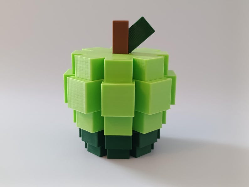 Puede incluir: Una escultura de manzana impresa en 3D, compuesta por cubos verdes apilados, con un tallo marr&oacute;n y una hoja verde oscuro. El cuerpo de la manzana transita de verde claro a oscuro, creando un efecto pixelado. Un objeto decorativo &uacute;nico.