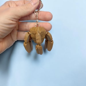 May include: A tan-colored, articulated crab keychain. The crab has a detailed, textured body and jointed legs. The keychain is attached to a silver metal ring and chain. The background is a solid light blue.