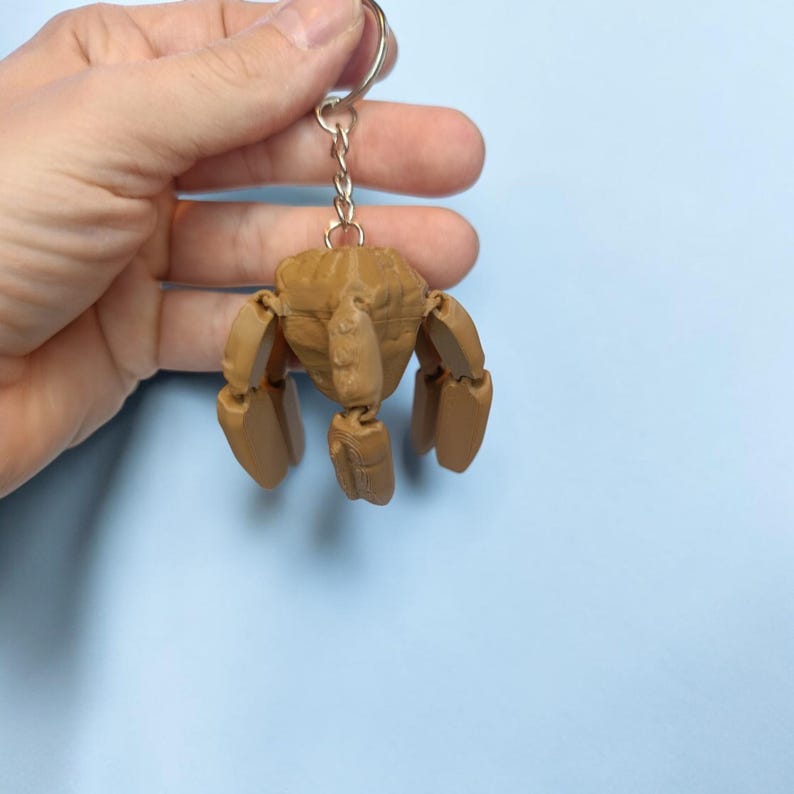 May include: A tan-colored, articulated crab keychain. The crab has a detailed, textured body and jointed legs. The keychain is attached to a silver metal ring and chain. The background is a solid light blue.