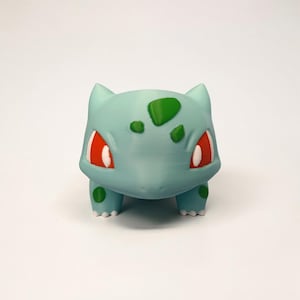 May include: A light blue Bulbasaur figurine with red eyes and green spots. The figurine has a rounded body, four small legs, and pointed ears. The design is a 3D-printed model of the popular Pok&eacute;mon character.