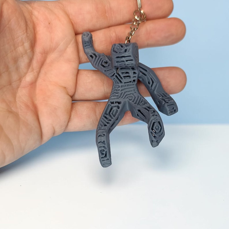 Puede incluir: Un llavero gris, impreso en 3D, con forma de figura humana estilizada. La figura tiene patrones geom&eacute;tricos y est&aacute; unida a un llavero de metal. El fondo es azul claro y blanco.