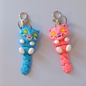 May include: Two articulated cat keyrings. One is blue with yellow eyes and white teeth, the other is pink with yellow eyes and white teeth. Both have silver-coloured metal clasps.