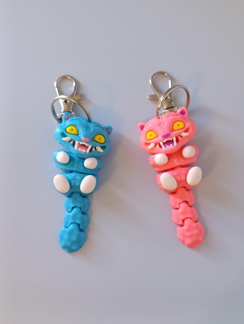 May include: Two articulated cat keyrings. One is blue with yellow eyes and white teeth, the other is pink with yellow eyes and white teeth. Both have silver-coloured metal clasps.
