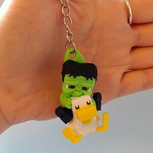 Op de afbeelding: Een groene Hulk-achtige figuur die een witte en gele eend-sleutelhanger vasthoudt. De sleutelhanger heeft een zilveren ketting en ring. De Hulk-achtige figuur heeft zwart haar en houdt de eend vast. De eend heeft een gele snavel en gele poten.
