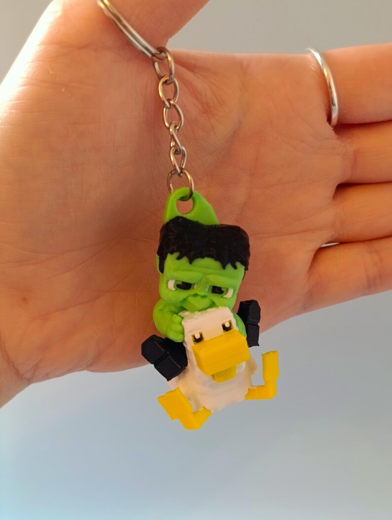 Op de afbeelding: Een groene Hulk-achtige figuur die een witte en gele eend-sleutelhanger vasthoudt. De sleutelhanger heeft een zilveren ketting en ring. De Hulk-achtige figuur heeft zwart haar en houdt de eend vast. De eend heeft een gele snavel en gele poten.