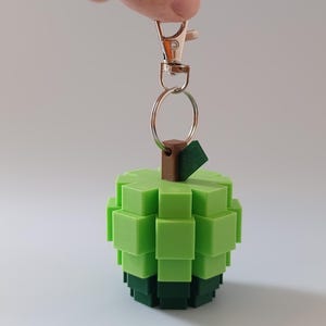 Puede incluir: Un llavero de manzana pixelada verde con un tallo marr&oacute;n y una base verde oscuro. El llavero est&aacute; sujeto a un mosquet&oacute;n y un anillo de metal plateado. La manzana est&aacute; hecha de peque&ntilde;os bloques cuadrados.