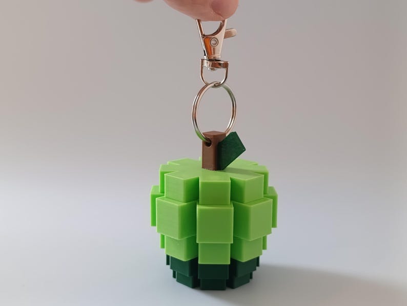 Puede incluir: Un llavero de manzana pixelada verde con un tallo marr&oacute;n y una base verde oscuro. El llavero est&aacute; sujeto a un mosquet&oacute;n y un anillo de metal plateado. La manzana est&aacute; hecha de peque&ntilde;os bloques cuadrados.