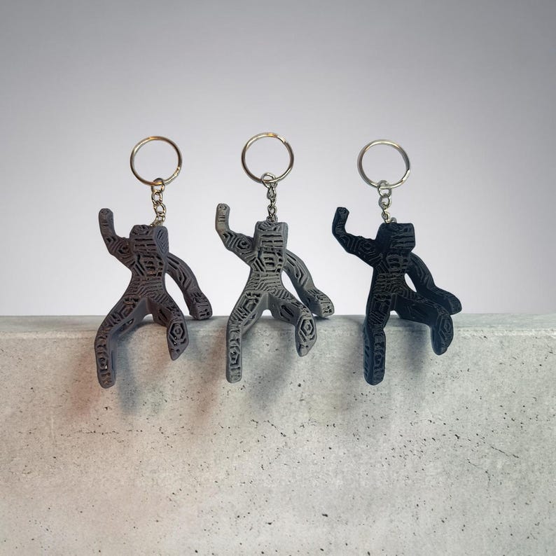 Puede incluir: Tres llaveros en fila, cada uno con una figura humana estilizada con los brazos extendidos. Los llaveros son en tonos grises y negros, con intrincados patrones grabados. Cada llavero tiene un anillo plateado.