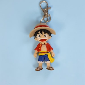 May include: A colorful keychain featuring a cartoon character with a straw hat, red jacket, blue shorts, and a yellow sash. The figure has a cheerful expression and is attached to a silver-colored metal clip.