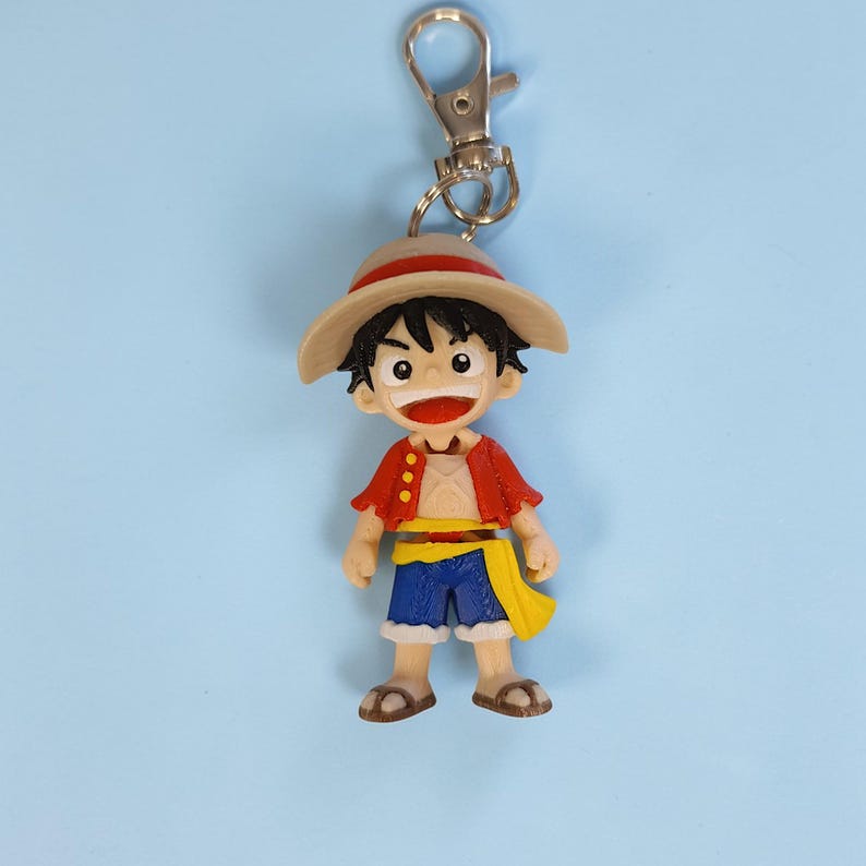 May include: A colorful keychain featuring a cartoon character with a straw hat, red jacket, blue shorts, and a yellow sash. The figure has a cheerful expression and is attached to a silver-colored metal clip.