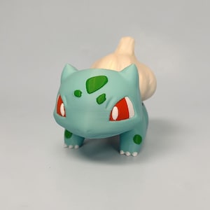May include: A 3D-printed Bulbasaur figure in a light teal color with green spots and red eyes. The figure has a bulb on its back. The design is based on the popular Pok&eacute;mon character.