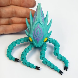 May include: A teal articulated creature toy with a spiky head and segmented tentacles. The head features purple accents and yellow eyes. The tentacles have black tips. The toy is designed to be poseable and is likely 3D printed.
