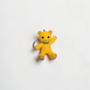Puede incluir: Un llavero de oso amarillo con una peque&ntilde;a pajarita de color verde azulado en el cuello, ojos negros circulares y un hocico de color marr&oacute;n claro. El oso aparece en una pose de saludo con un brazo levantado e incluye una anilla de metal plateado.