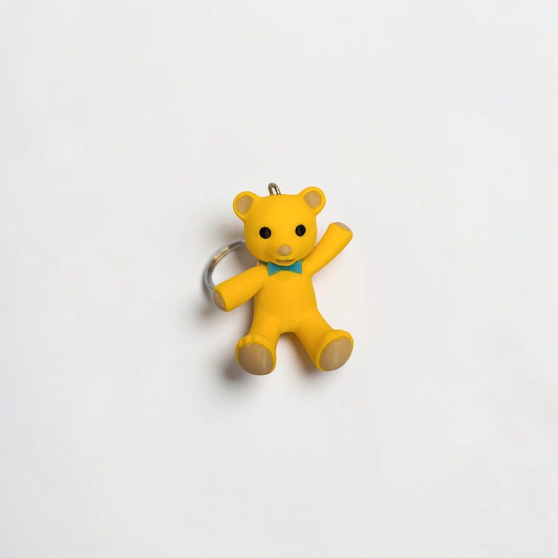 Puede incluir: Un llavero de oso amarillo con una peque&ntilde;a pajarita de color verde azulado en el cuello, ojos negros circulares y un hocico de color marr&oacute;n claro. El oso aparece en una pose de saludo con un brazo levantado e incluye una anilla de metal plateado.