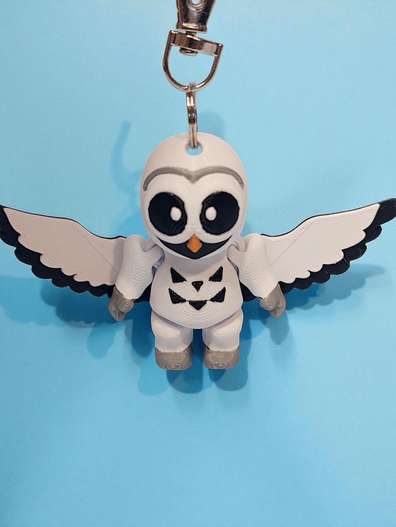 May include: A white owl-shaped keychain with black eyes, a gray beak, and gray accents. The owl has outstretched wings with black tips and gray feet. A silver-colored metal clasp is attached to the top of the owl's head.