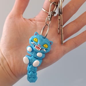 May include: A blue cat-shaped keyring with yellow eyes and white teeth. The keyring is attached to a silver clasp and ring, with several keys hanging from the ring. The cat has a segmented body and white paws.